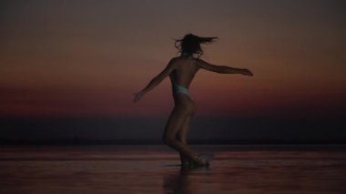 Hafif mayo giymiş bir kadın sığ sularda zarifçe hareket eder, saçları gün batımının derin renkleri gibi kıvrılır dramatik ve sakin bir zemin oluşturur.. 