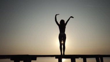 Bikinili siluetli bir kadın, ahşap bir iskelede zarif bir şekilde duruyor. Sıcak günbatımının ışığı aşağıdaki sakin suları yıkarken kolları narin bir pozda yükseliyor.. 