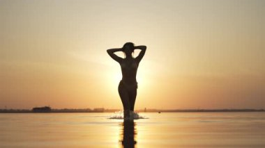 Bir kadın sığ suda yürüyor, kolları zarif bir şekilde başının arkasında, silueti günbatımının altın parıltısıyla güzelce çizilmiş.. 