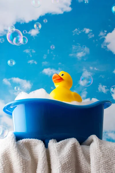 Rubber yellow duck sits in a doll bath with soap suds, children\'s spa and hygiene concept, children\'s bathing accessories on a background of blue sky and soap bubbles