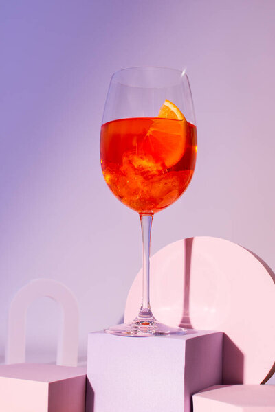 A glass with an alcoholic cocktail on a podium, an aperol with dry wine and ice and a slice of orange close-up in delicate pastel colors