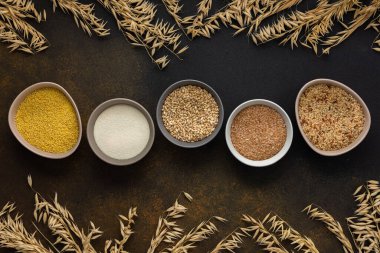 Kâselerdeki tahıl gevrekleri, kahverengi bir arka planda mısır gevreği kaseleri ve yulaf kulakları olan üst manzara.