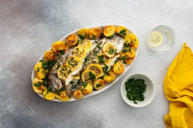 Oven-baked Dorada fish with potatoes and lemon slices, sprinkled with lemon juice and mashed parsley, cooked fish in an oval dish, top view