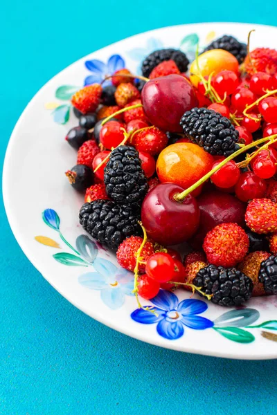 Fresh berries on a plate on a turquoise background, red and black currants and black mulberries, wild strawberries and cherries, top view