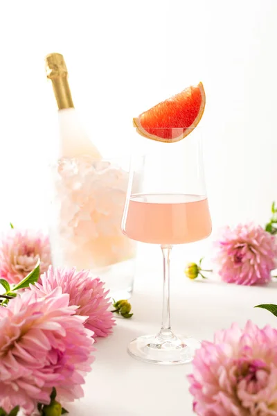 A glass with rose sparkling wine surrounded by dahlias, a bottle of rose sparkling wine is chilled in a glass bucket with ice , grapefruit slice in a glass