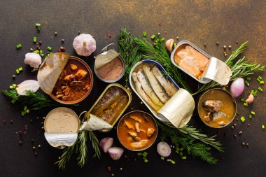 Baharat ve bitkilerin arasında farklı teneke kutular, kahverengi bir arka planda konserve somon ve uskumru, sprat ve sardalya, ton balığı ve ringa balığı ve balık ezmesi.