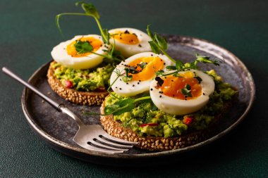 Çavdar ekmekli sandviç ve guacamole, haşlanmış yumurta, bezelye ve turp ve susam tohumlarıyla birlikte, yeşil arka planda yakın plan sandviçli bir tabak.