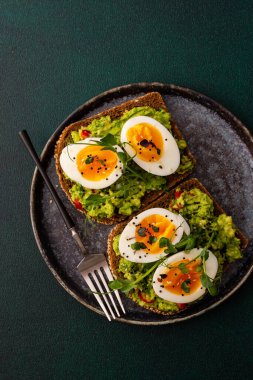 Çavdar ekmekli sandviç ve guacamole, haşlanmış yumurta, bezelye ve turp ve susam tohumlarıyla birlikte, yeşil arka planda yakın plan sandviçli bir tabak.