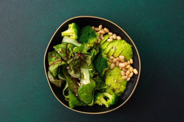 Sağlıklı kahvaltı, fasulye, salatalık, avokado ve brokoli serpiştirilmiş yeşil arka planda bir tabak yemek.