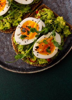 Çavdar ekmekli sandviç ve guacamole, haşlanmış yumurta, bezelye ve turp ve susam tohumlarıyla birlikte, yeşil arka planda yakın plan sandviçli bir tabak.