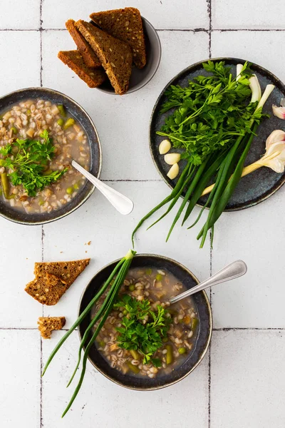 Healthy lunches, soup with pearl barley and asparagus and green peas, mushrooms and potatoes, top view of bowls of soup and a plate with green onions, garlic and parsley, rye bread