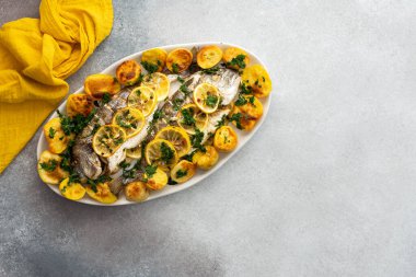 Oven-baked Dorada fish with potatoes and lemon slices, sprinkled with lemon juice and mashed parsley, cooked fish in an oval dish, top view, copy space