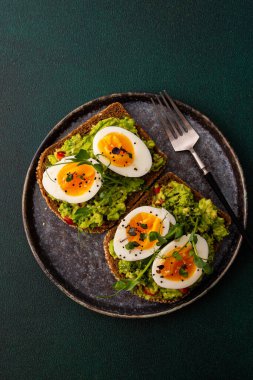 Çavdar ekmekli sandviç ve guacamole, haşlanmış yumurta, bezelye ve turp ve susam tohumlarıyla birlikte, yeşil arka planda yakın plan sandviçli bir tabak.