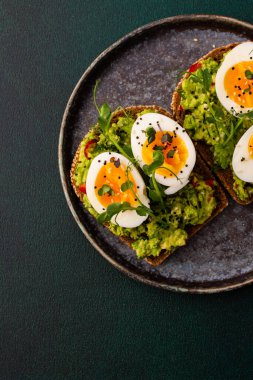 Çavdar ekmekli sandviç ve guacamole, haşlanmış yumurta, bezelye ve turp ve susam tohumlarıyla birlikte, yeşil arka planda yakın plan sandviçli bir tabak.