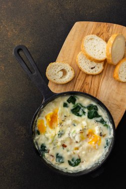 Sağlıklı kahvaltı veya akşam yemeği, peynirli sahanda yumurta, ıspanak yaprağı ve hindistan cevizi sütü dökme demir tavada, kahverengi arka planda dilimlenmiş baget.