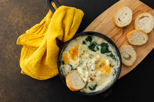 Healthy breakfasts or dinners, fried eggs with cheese, spinach leaves and coconut milk in a cast-iron pan, sliced baguette on a cutting board on a brown background