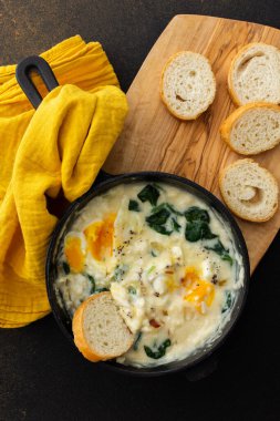 Sağlıklı kahvaltı veya akşam yemeği, peynirli sahanda yumurta, ıspanak yaprağı ve hindistan cevizi sütü dökme demir tavada, kahverengi arka planda dilimlenmiş baget.