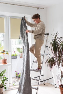 Bir kadın merdiven üzerinde durur ve gri perdeleri dairedeki bir korkuluğa asar, bir apartmanda bahar temizliği yapar, perdeleri diker ve onları asmak için hizmet verir.