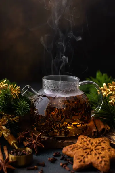 Glass teapot with brewed black tea with steam among fir branches and golden Christmas decorations, gingerbread and spices, cozy festive winter evening