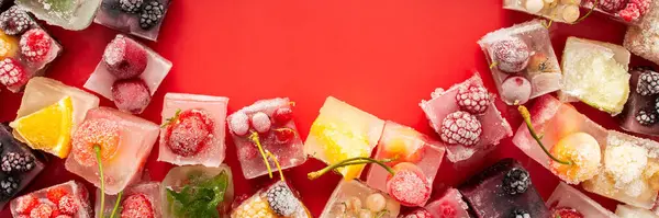 Frame of ice cubes with frozen berries and fruits and mint on red background, banner