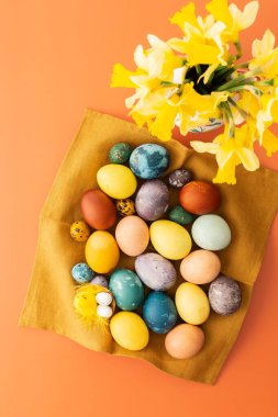 Doğal boyalı peçetenin üst görüntüsü Paskalya yumurtaları, turuncu arka planda vazoda sarı nergis çiçekleri.
