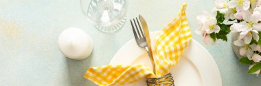A charming table setting for spring includes a white plate with a yellow checkered napkin, fork, and knife neatly arranged among colorful flowers in bloom, banner