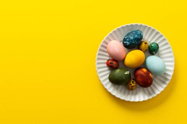 A variety of beautifully painted Easter eggs in different colors and patterns are placed on a delicate white plate. The vibrant hues stand out against the cheerful yellow backdrop.