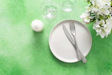 A lovely spring table setting features a white plate, silverware, and elegant glassware, complemented by fresh flowers and a soft green background, perfect for gatherings and meals.