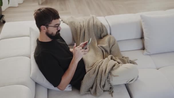 남자는 편안하게 소파에 자신의 전화와 재미있는 순간을 공유하고있다 허벅지는 안경을 — 비디오