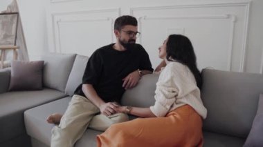 Bir erkek ve bir kadın rahat bir şekilde kanepede otururlar, el ele tutuşup, birbirlerine derin derin bakarlar, başparmaklarını kenetlerler.