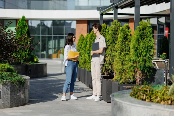 Laptoplu genç erkek ve kitaplı kadın öğrencilerin yan görüntüsü. Sokakta durmuş kampüs binasına karşı yeşil ağaçlarla tartışırken birbirlerine bakıyorlar. 