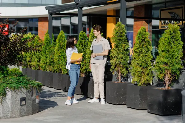 Laptoplu genç erkek ve kitaplı kadın öğrencilerin yan görüntüsü. Sokakta durmuş kampüs binasına karşı yeşil ağaçlarla tartışırken birbirlerine bakıyorlar. 