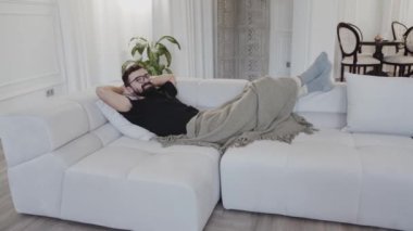 Saf ahşap döşemeleri olan, iç dekorasyonda mobilya ve bitkilerle çevrili, rahat bir oturma odasında beyaz bir kanepeye yaslanmış bir adam var.