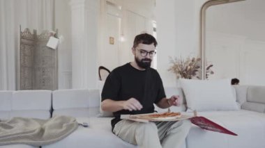 Sakallı bir adam bir odada bir kanepede oturuyor ve elinde bir tepsi pizza var.
