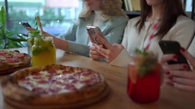 Bir grup kadın bir etkinlik sırasında telefonlarına bakarken bir masada pizza ve içki paylaşıyorlar.