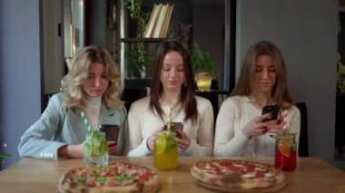 Üç kadın, pizza, içki ve sofra takımı ile bir masanın etrafında toplanıyor, bir evde yemek yerken telefonlarına sarılıyorlar.