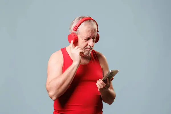 Elinde kulaklıklarla akıllı telefonunu kullanan son sınıf öğrencisi, bu stüdyoda mavi bir arkaplanda çekim yapıyor. Yaşlılar için teknoloji kullanımı, müzik dinlemek ve uygulamalar kullanmak..