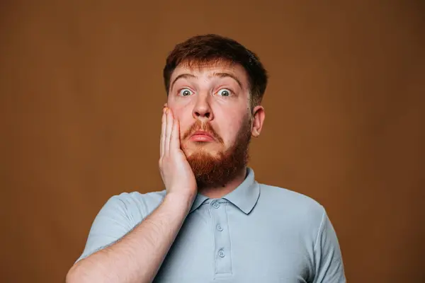 Sakallı bir adam elini yüzüne tutuyor, kolu sakalına sürtünürken yanağına koluyla dokunuyor. Bu jest, onun gömleğinin çenesine ve yakasına dikkat çekiyor.