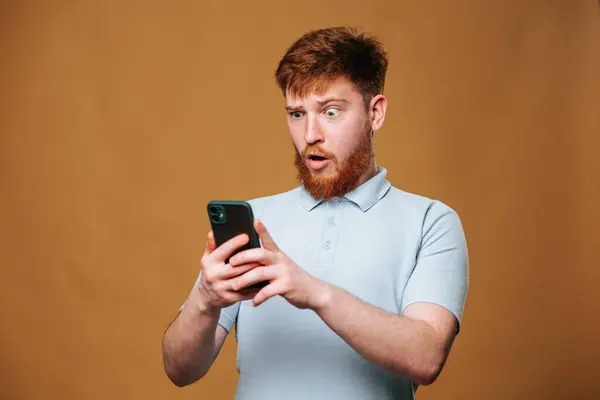 A teenage redhead guy in a studio shot, shocked and staring at his smartphone with a scared expression on his face, An adolescent guy with a shocked expression on his face, looking at his smartphone