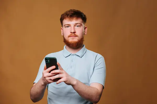 Yüzü kıllı bir adam başparmağı sakalının yanındayken elinde bir cep telefonu tutuyor. Gözlük takıyor ve dirseği de kolunda dinleniyor.