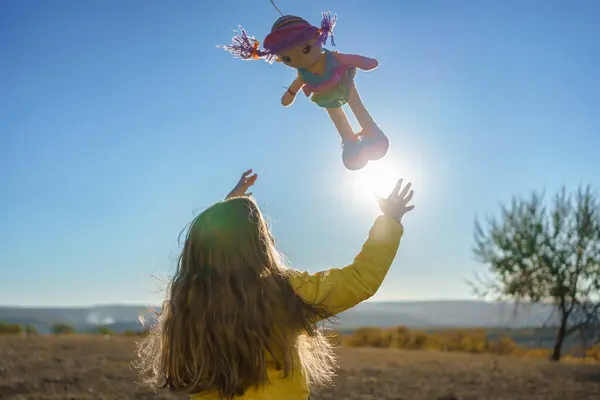 Genç kız dışarıda oynuyor, oyuncak bebeği gökyüzüne fırlatıyor, güneşli bir günde kaygısız neşe ve masumiyeti somutlaştırıyor.
