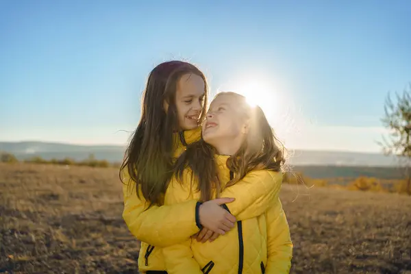Güneşli bir açık hava ortamında, sarı ceketli iki genç kız birbirlerine sarılıp neşeli bir anı paylaşıyorlar.