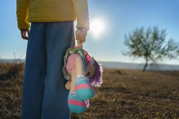 Sarı paltolu bir çocuk oyuncak bir at tutuyor. Gün doğumunda bir tarlada duruyor. Uzaktaki bir ağaçla..