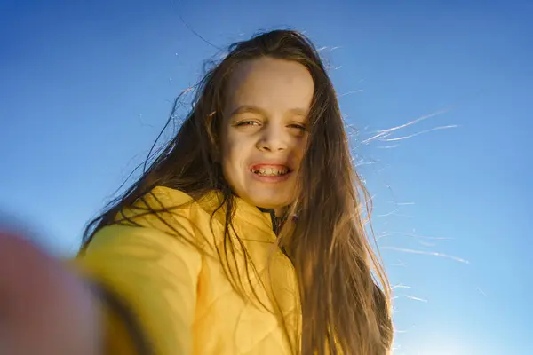Sarı ceketli, uzun saçlı genç bir kız mavi gökyüzünün altında selfie çekiyor, neşe ve özgürlüğü yakalıyor.