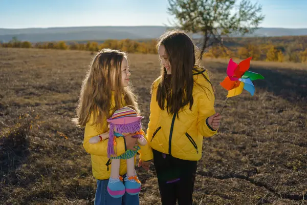 Sarı ceketli, oyuncak bebekli ve fırıldaklı iki kız kuru bir tarlada güneşli bir günün tadını çıkarıyorlar.