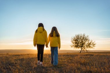Sarı ceketli iki kız gün batımında bir tarlada el ele yürür, dostluğu ve huzuru simgeler..