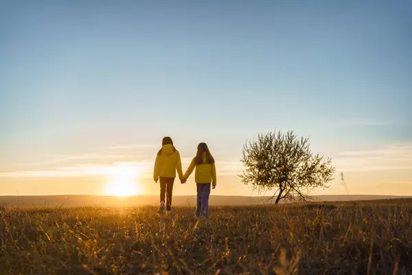 Sarı ceketli iki çocuk, gün batımına doğru yalnız bir ağaçla yürüyorlar..