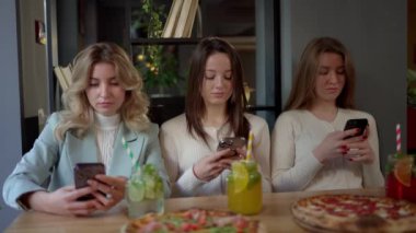 Üç kadın pizza ve içeceklerle bir masanın etrafında toplanıyor, telefonlarına bakarken gülücüklerini paylaşıyorlar. Bu sakin etkinlik bir binada gerçekleşiyor ve toplantılarına biraz eğlence katıyor.
