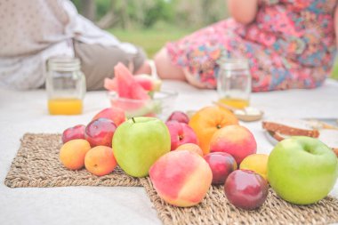 Unrecognizable couple having a picnic breakfast with a variety of fresh fruits, apples, peaches, tangerines, plums, watermelon. Focus on them. Concepts: healthy eating, enjoying the outdoors at summer