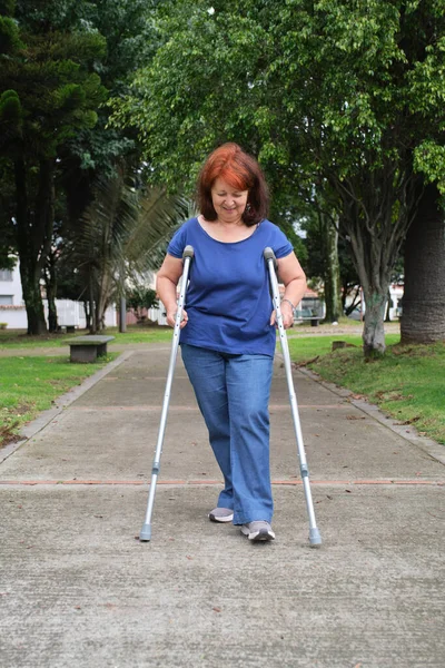 Mature hispanic human recovering from an injury walking with crutches on an urban park pathway, smiling. Concepts: positive attitude towards adversity, physical rehabilitation